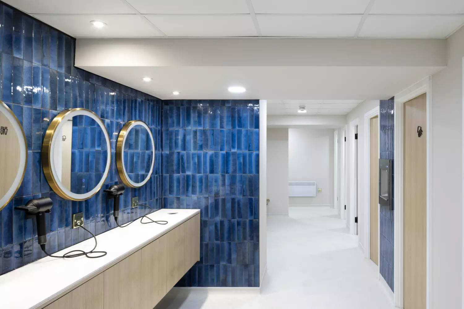 A modern changing area with blue tiled walls, round gold framed mirrors and wall mounted hairdryers. A hallway leads to several doors, likely for individual stalls or rooms. Recessed ceiling lights and light coloured tiled flooring contribute to the clean, functional design.